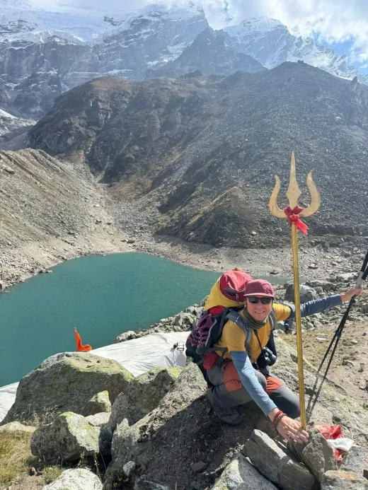 Martin kneeling by a trident and prayer flags above a turquoise glacial lake in the Himalaya