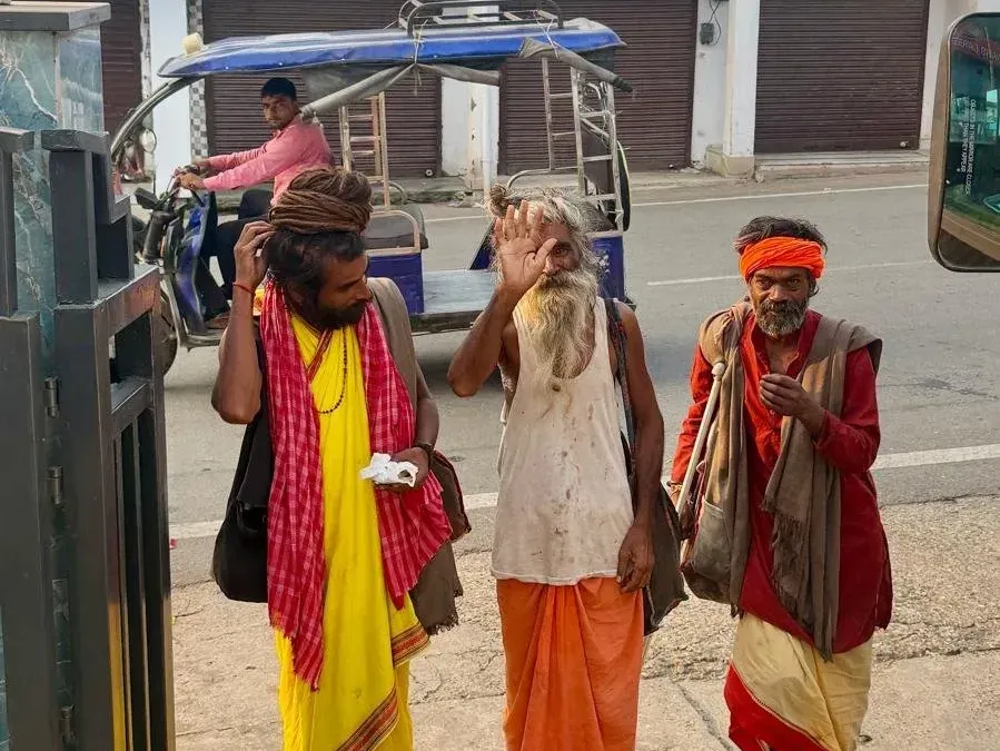 Three sadhus in yellow, saffron and red robes standing by a street, one raising his hand in greeting