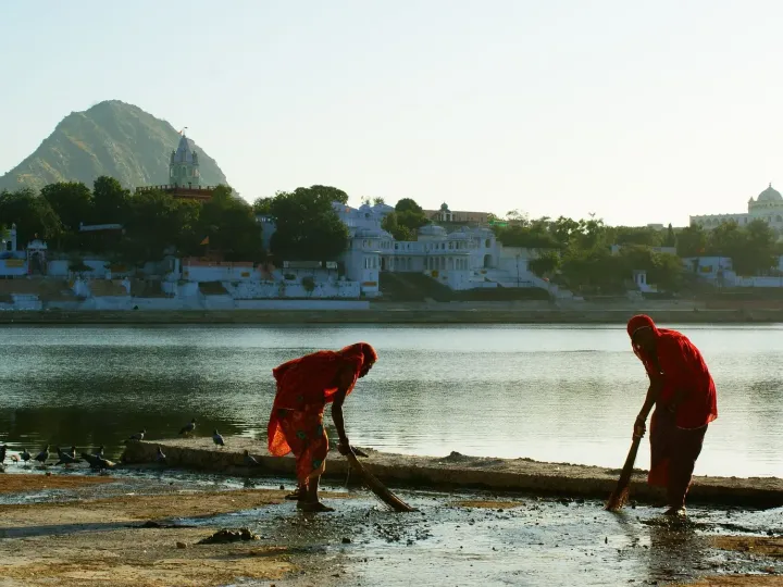 Women sweeping the ghats of the sacred lake in Pushkar at sunrise, with temples and a hill behind